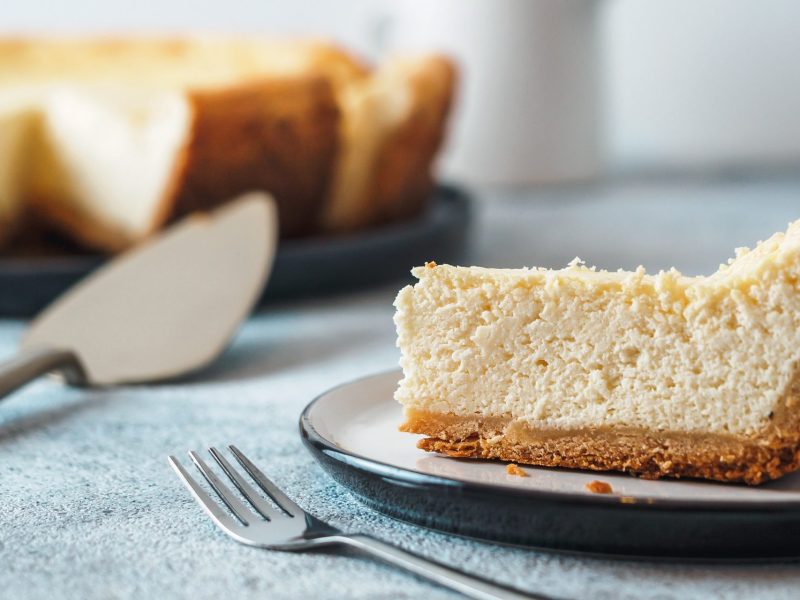 Ein Stück Mango-Vanille-Cheesecake auf einem Teller, daneben eine Gabel, im Hintergrund der angeschnittene Kuchen.