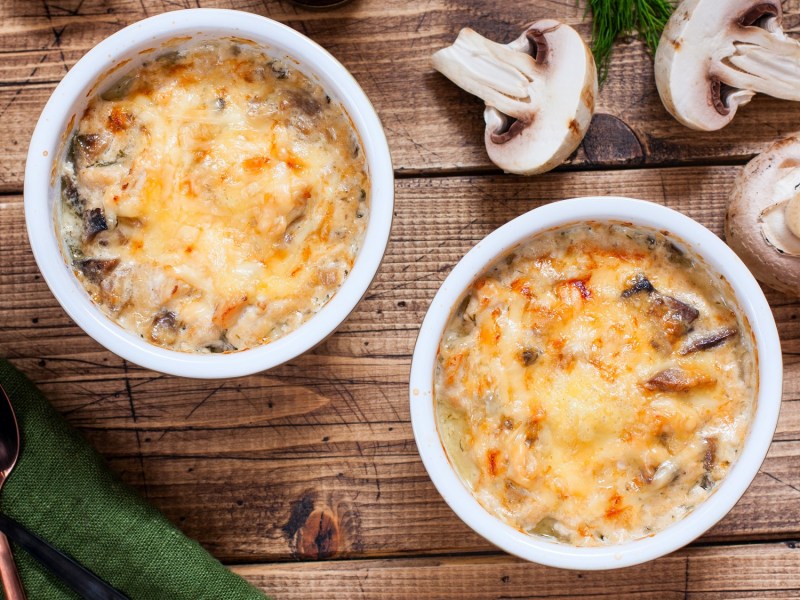 Draufsicht: Zwei runde Auflaufformen einem Penne-Champignon-Auflauf stehen auf einem Holztisch. Drum herum liegen frische Pilze.