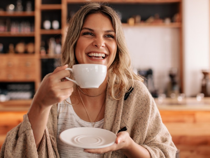 Frau mit blonden Haaren lacht und hält Kaffeetasse in Hand.