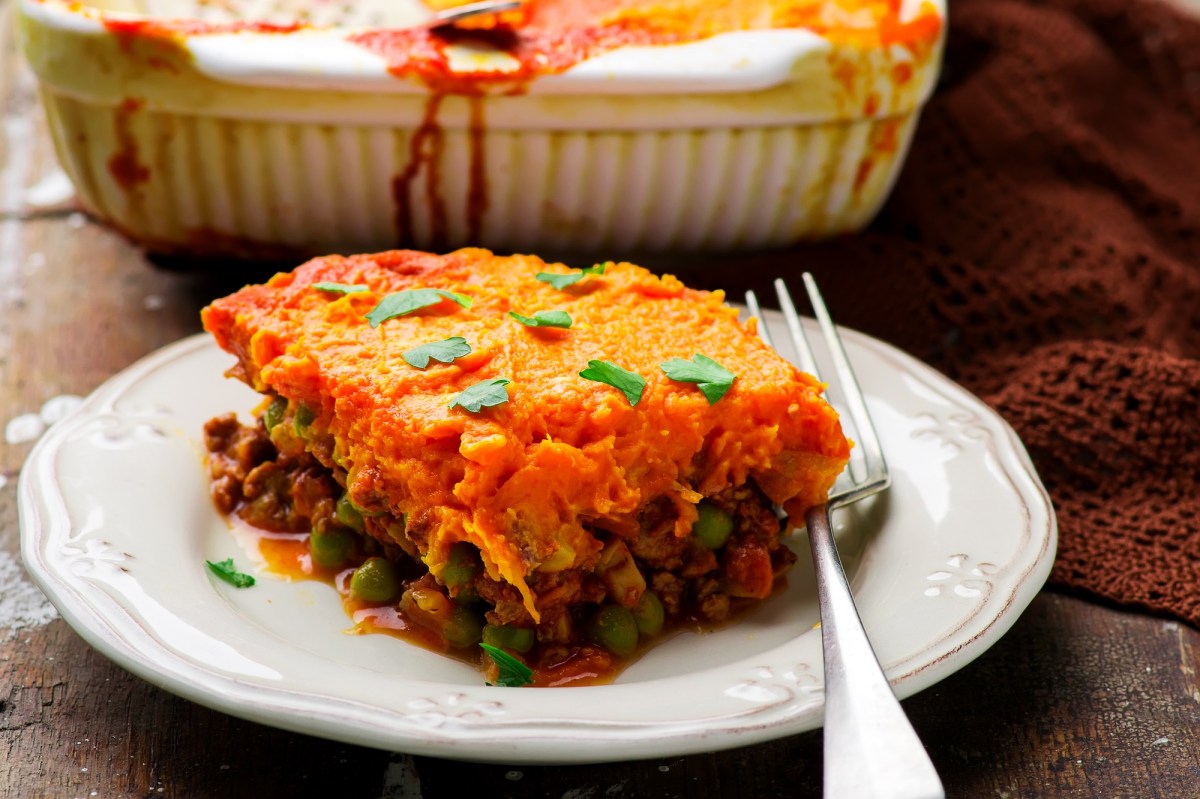 Ein Stück Shepherd's Pie mit Süßkartoffelbrei auf einem Teller, daneben eine Gabel, dahinter die Auflaufform.