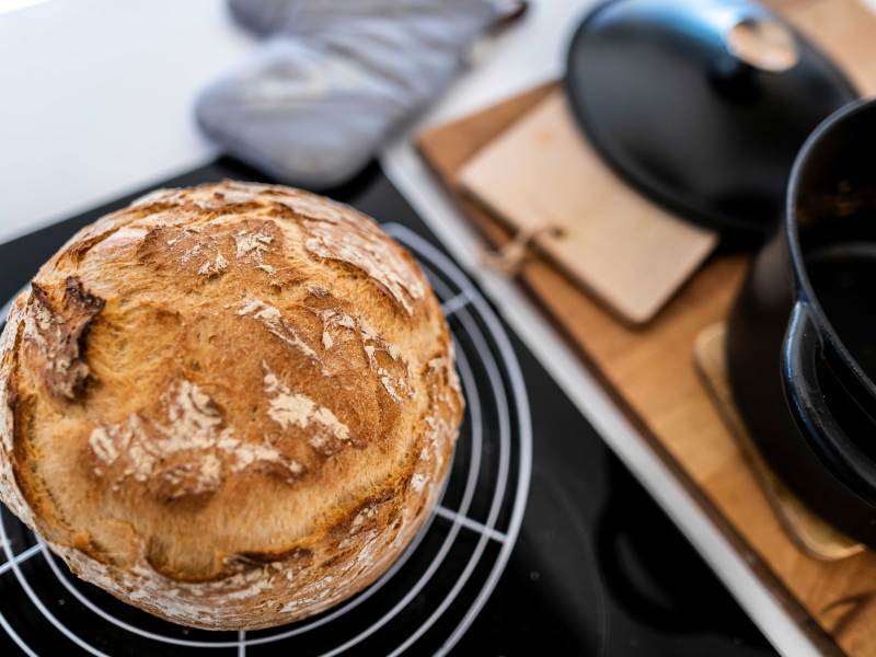 Brotlaib kühlt auf einem Küchenrost ab, daneben steht noch der Topf, in dem es im Ofen gebacken wurde.