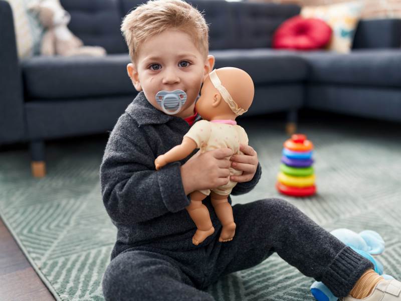 Ca. dreijähriger Junge sitzt mit einer Puppe im Arm und einem Nuckel im Mund im Zimmer und spielt.