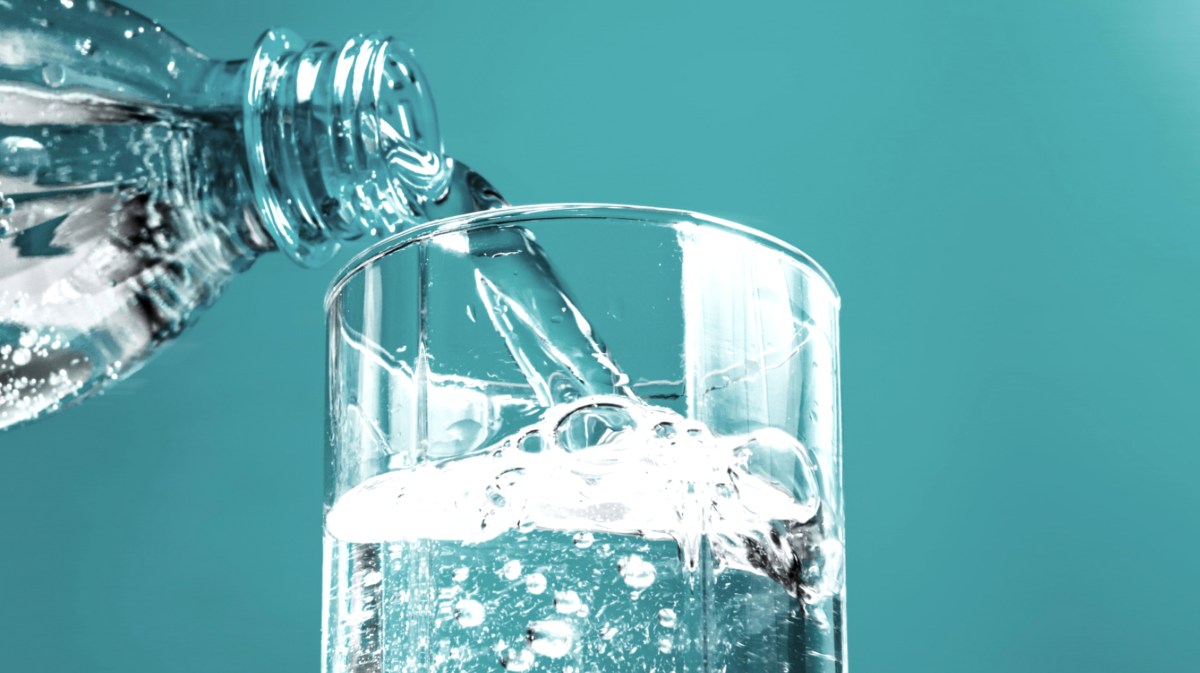 Mineralwasser wird aus einer Glasflasche in ein Glas geschüttet. Der Hintergrund ist türkis.
