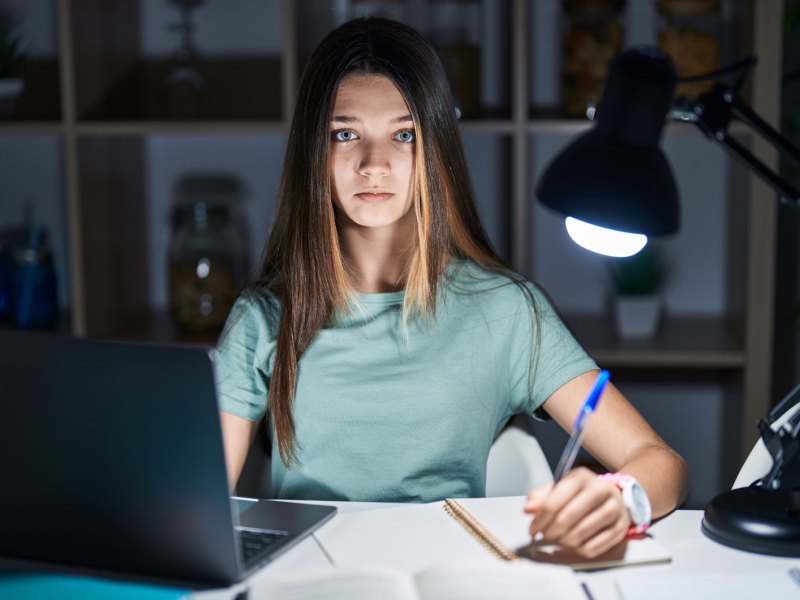 Teenagermädchen sitzt im Dunkeln an ihrem Schreibtisch, der von einer Schreibtischlampe beleuchtet wird und lernt.