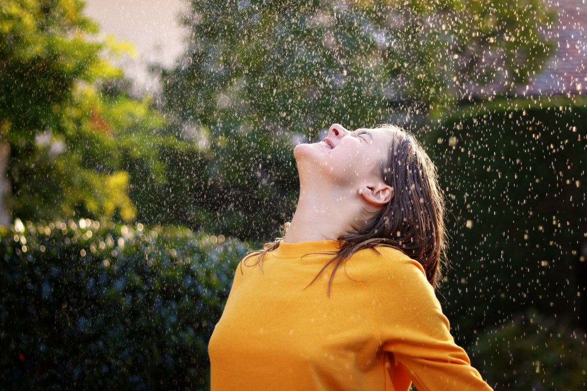 Frau steht im Sommerregen und streckt lächelnd das Gesicht zum Himmel.