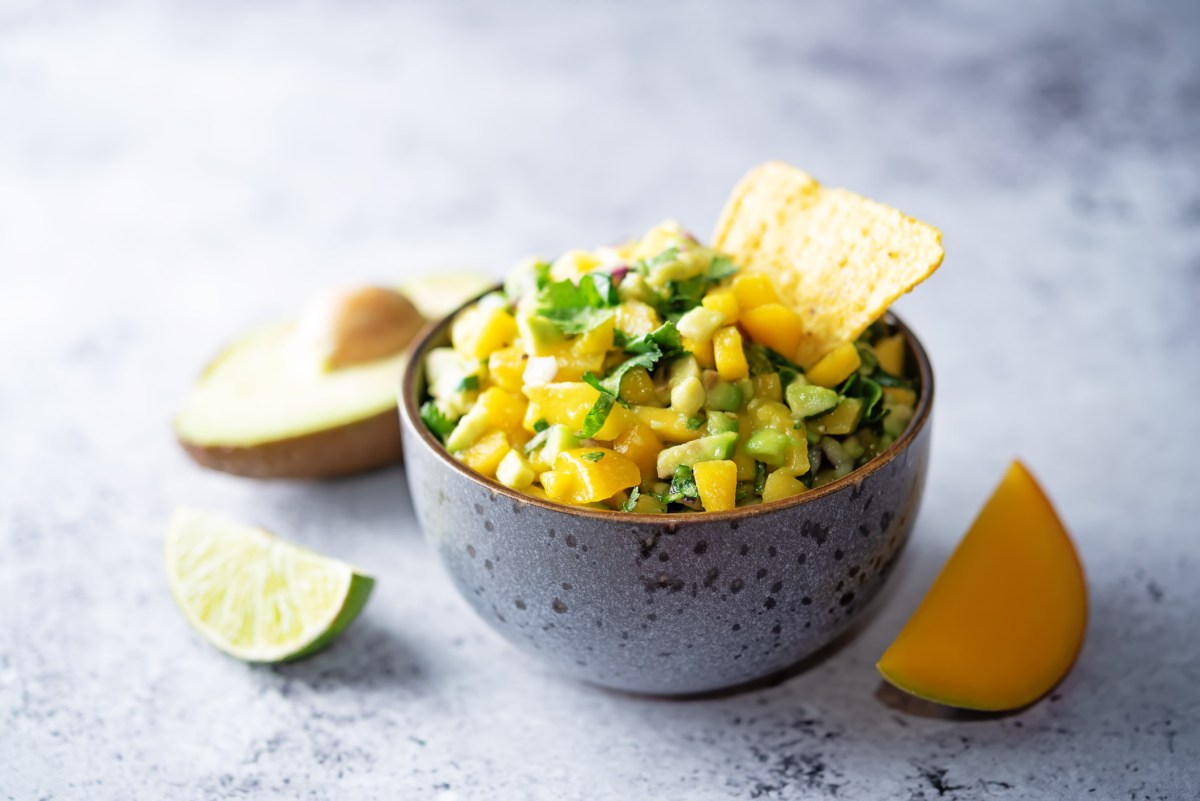 Eine Schüssel Mango-Avocado-Salsa mit eine Nacho-Chip, daneben je eine angeschnittene Limette, Avocado und Mango.