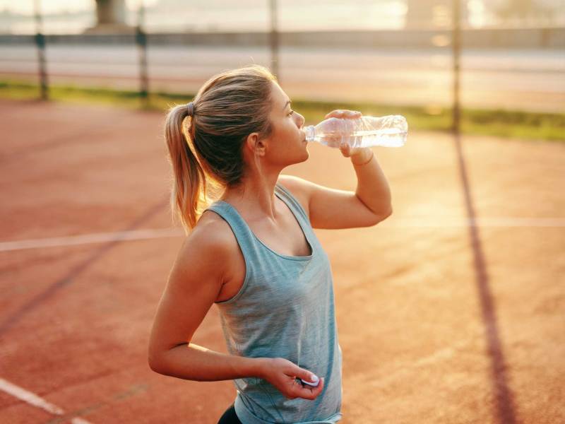 Sportliche blonde Frau steht auf dem Platz und trinkt Wasser nach dem Morgensport.