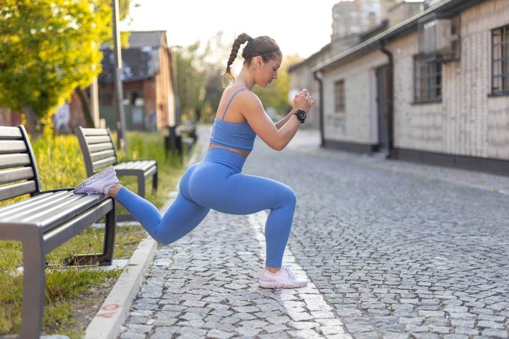 Junges weibliches Fitnessmodell in blauer Trainingskleidung macht einbeinige Split-Squats auf einer Bank.
