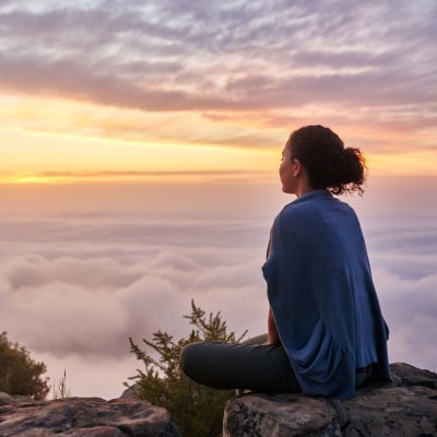 A woman on a mountain sits cross-legged on a rock and watches the sunrise above the clouds