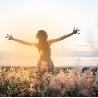 A woman in a field of flowers among the sun's rays raises her arms forcefully to the side
