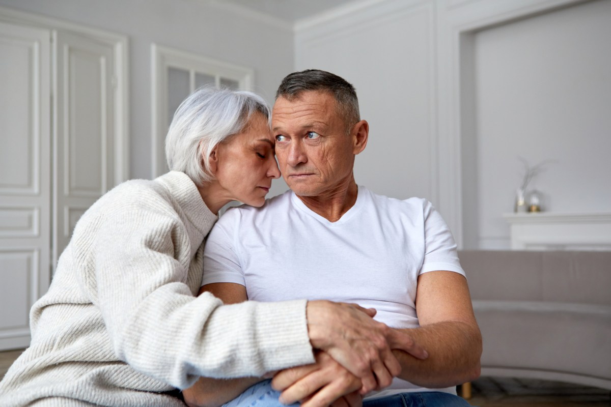 Älteres Paar mit grauen Haaren sitzt unglücklich auf einem Sofa. Sie hat die Arme um ihn geschlungen.