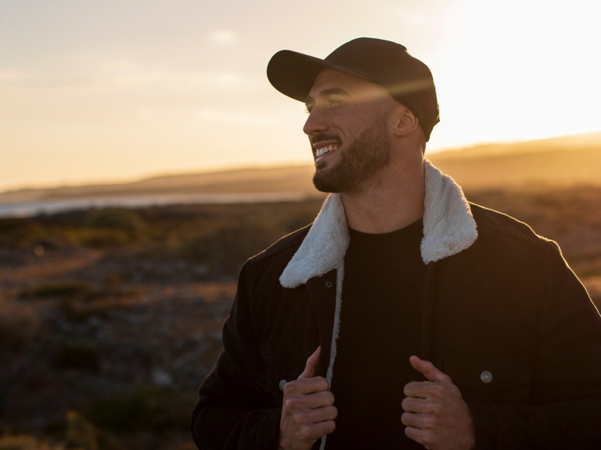 Mann in der Natur beim Sonnenuntergang, der seine Hände zur Jacke nimmt und lächelnd zur Seite schaut