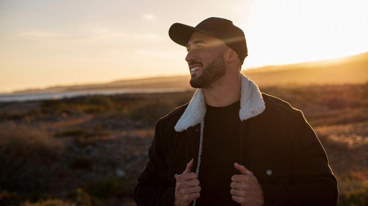 Mann in der Natur beim Sonnenuntergang, der seine Hände zur Jacke nimmt und lächelnd zur Seite schaut