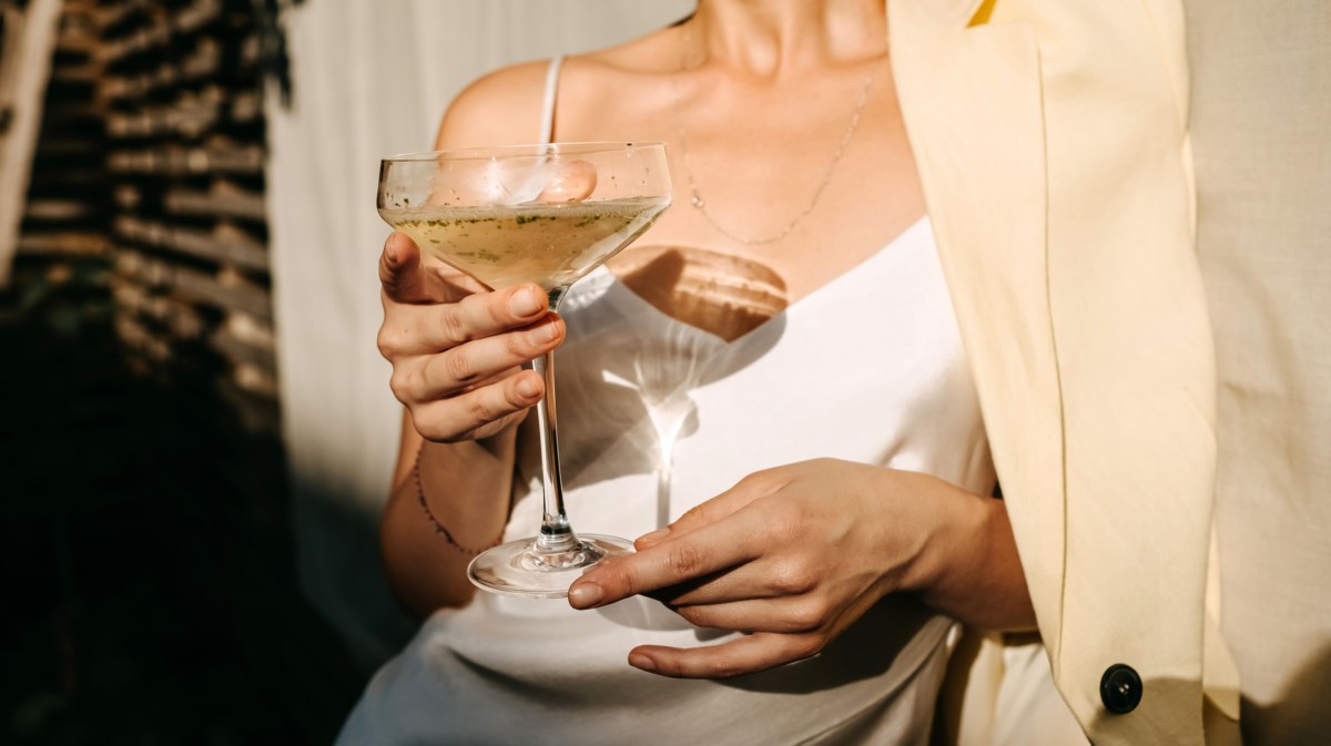 Frau in elegantem weißen Kleid, die ein Cocktailglas in der Hand hält und von der Sonne bestrahlt wird