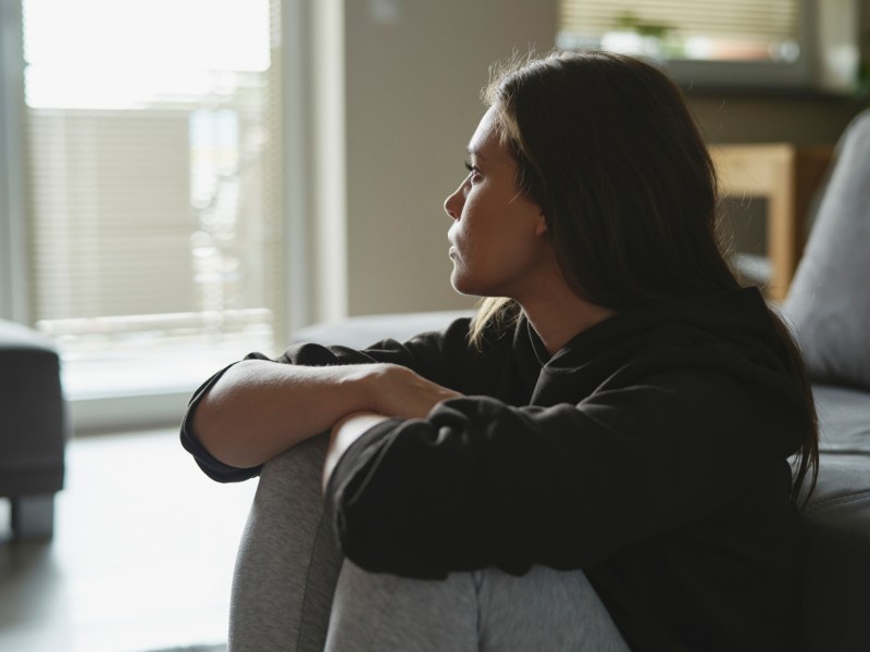Frau schaut nachdenkllich und traurig aus Fenster, während sie auf Holzboden vor Couch sitzt