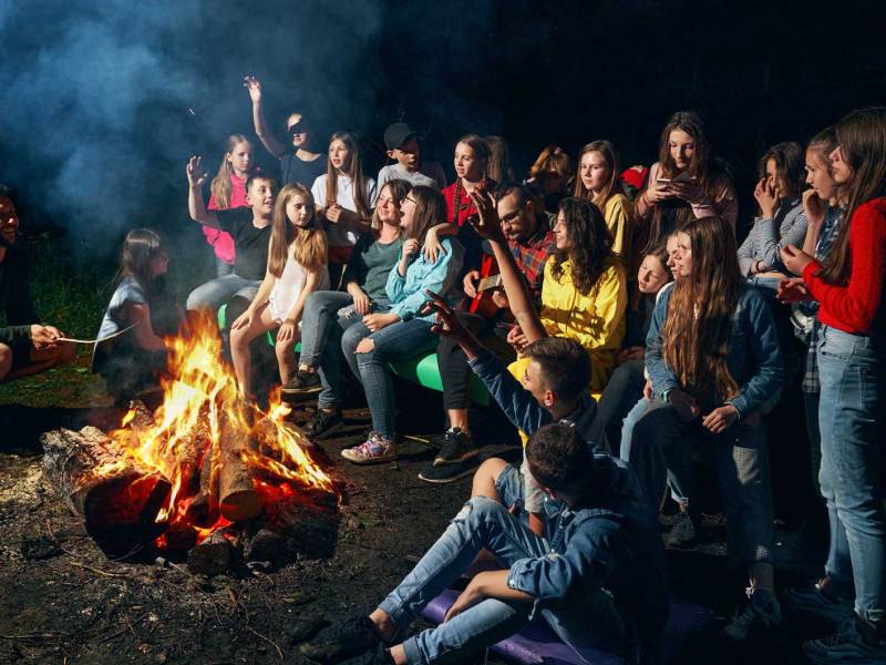 Zahlreiche Kinder sitzen und stehen um ein Lagerfeuer herum. Vor dem Feuer hockt auch ein Erwachsener.