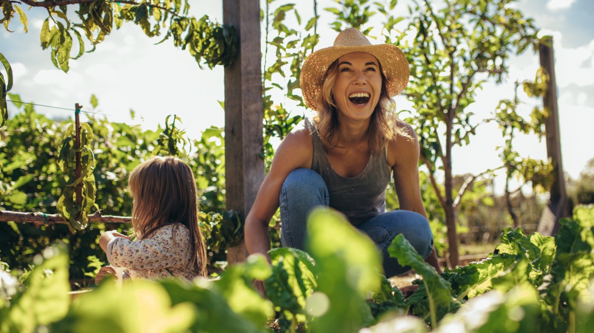 Frau mit Hut auf dem Kopf die in einem Garten voller Pflanzen steht und in der Sonne lacht