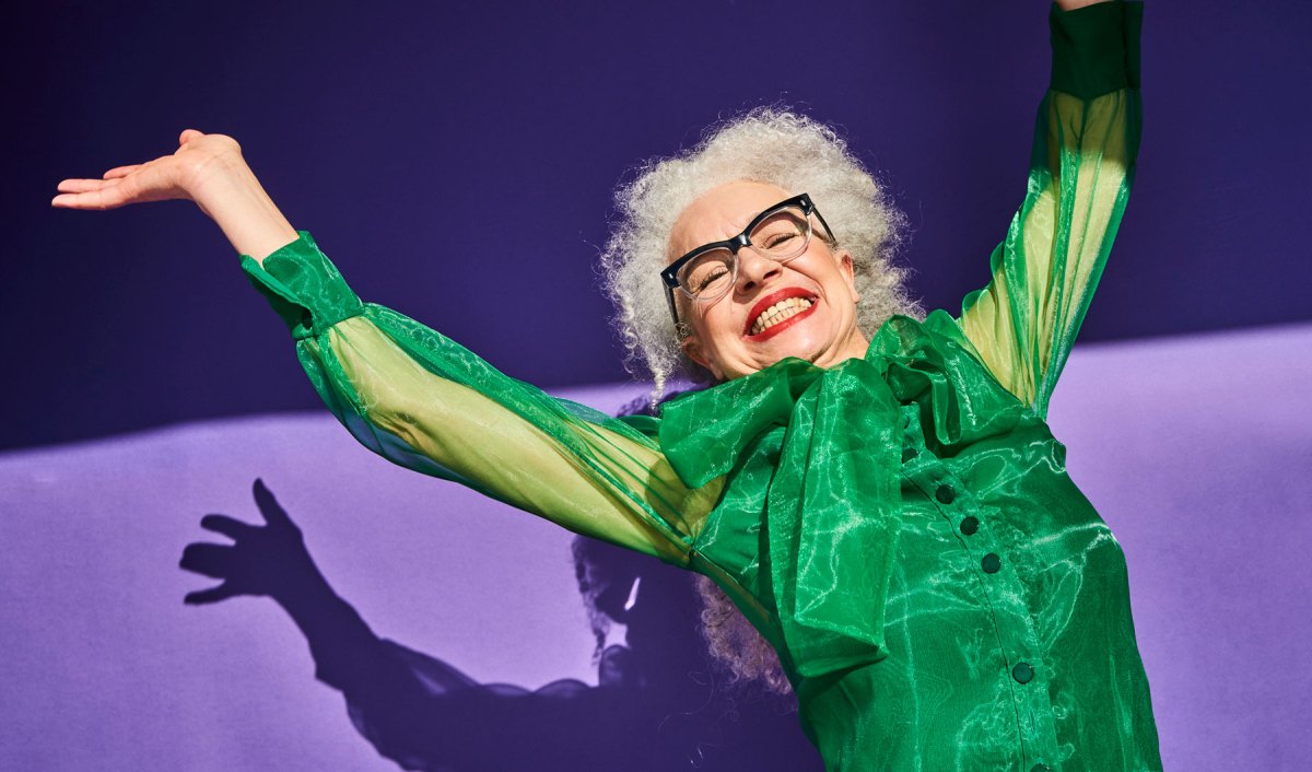 Ältere Frau mit grünem Kleid tanzt ausgelassen vor lila Hintergund