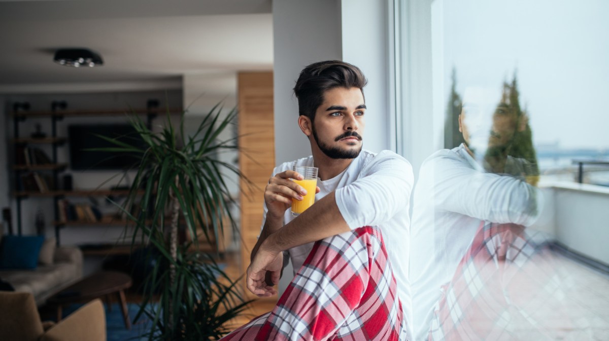 Mann, der mit Schlafanzug und Kaffee-Tasse in der Hand am Fenster sitzt und nachdenklich herausschaut