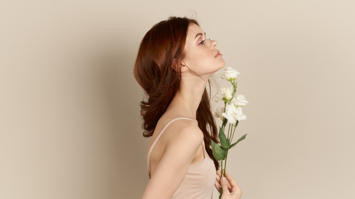 Rothaarige Frau mit weißen Blumen in der Hand, die zur Seite steht und nach oben blickt, während sie vor einer beigen Wand steht