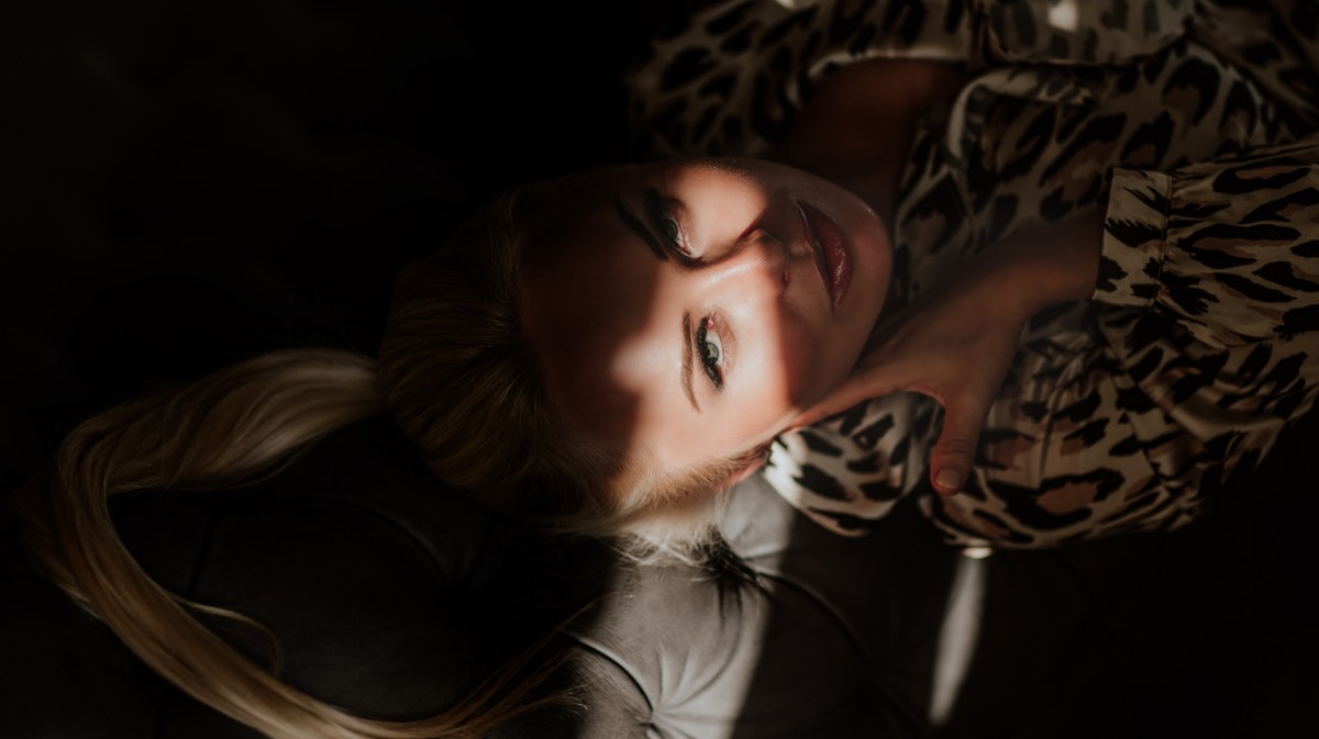 Frau auf einer dunklen Couch, die einen Sonnenstrahl zwischen ihrem Gesicht hat und mit tiefem Blick in die Kamera schaut