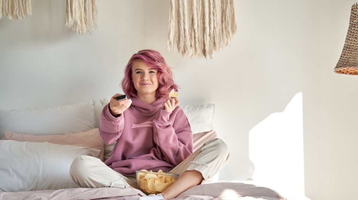 Frau mit rosa Haaren auf ihren Bett, die Chips isst und eine Fernbedienung in der Hand hält