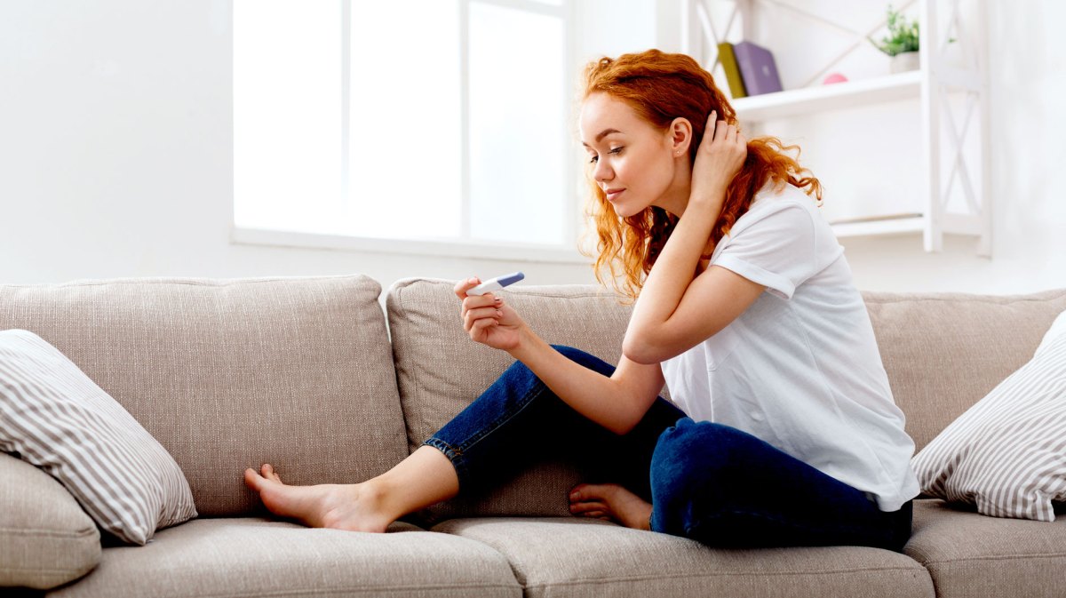 Junge Frau sitzt auf dem Sofa und schaut erwartungsvoll auf einen Schwangerschaftstest in ihrer rechten Hand.