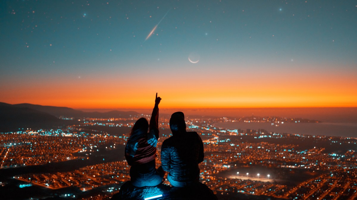 Mann und Frau im Sonnenuntergang auf einem Fels vor einer leuchtenden Stadt, während die Frau mit dem Finger auf eine Sternschnuppe zeigt