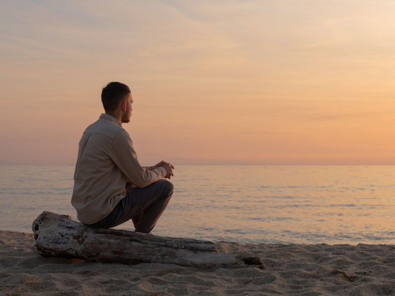Mann am Strand, der mit dem Rücken zur Kamera sitzt und auf das Meer schaut im Sonnenuntergang