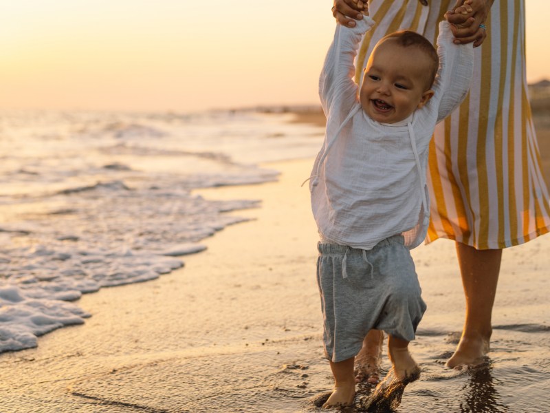 Frau mit Baby am Strand, das durch den Sand am Wasser geht und sich mit den Armen an der Mutter festhält im Sonnenuntergang