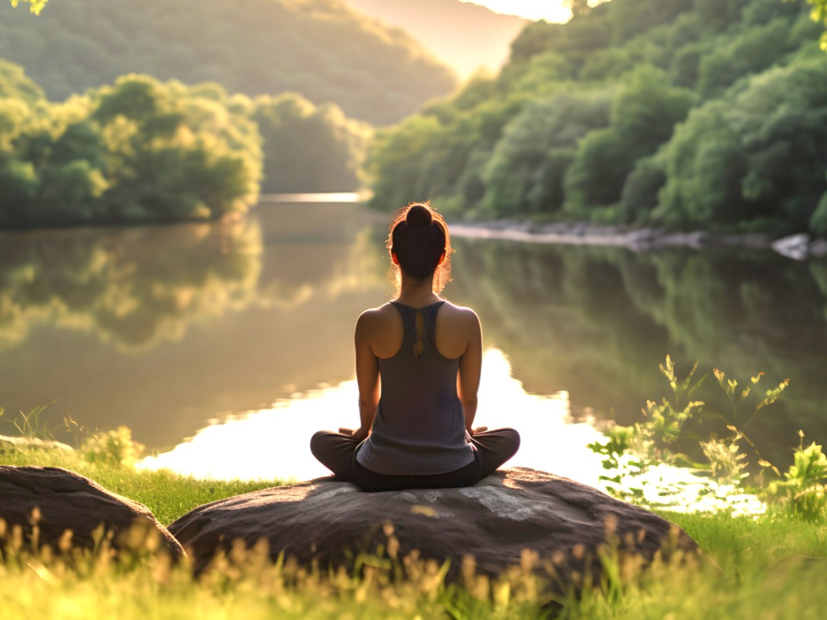 Eine Frau sitzt auf einem Stein am See und meditiert.