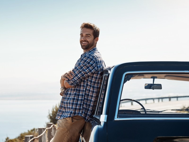 Mann mit kariertem Hemd und hellbrauner Hose lehnt sich an ein Auto und schaut zur Seite, während er auf einem Berg steht, wo im Hintergrund Wasser zu sehen ist
