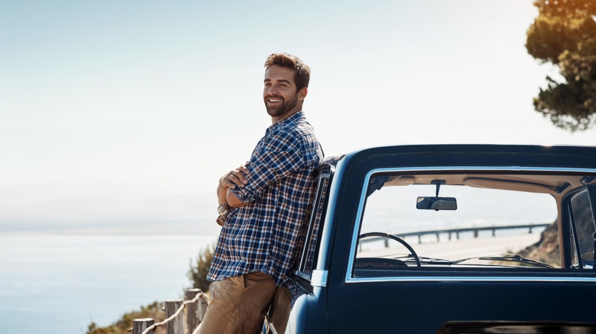Mann mit kariertem Hemd und hellbrauner Hose lehnt sich an ein Auto und schaut zur Seite, während er auf einem Berg steht, wo im Hintergrund Wasser zu sehen ist