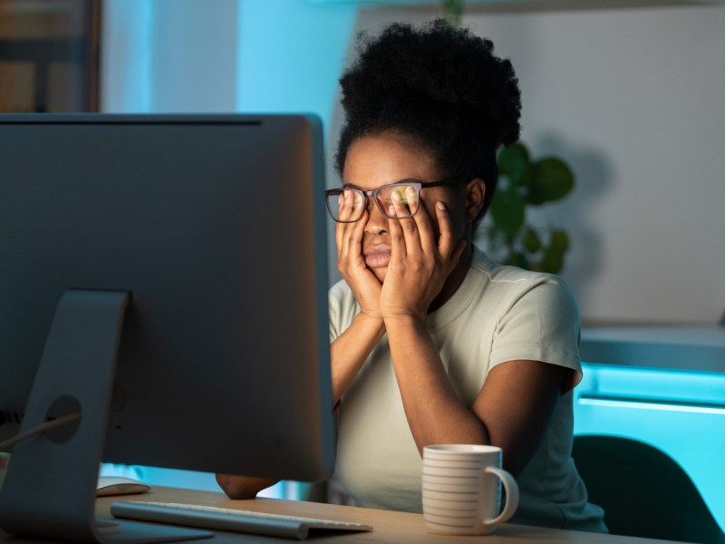 Frau sitzt mit Brille im Dunkeln vor ihrem PC Bildschirm und reibt sich verschlafen und überfordert die Augen