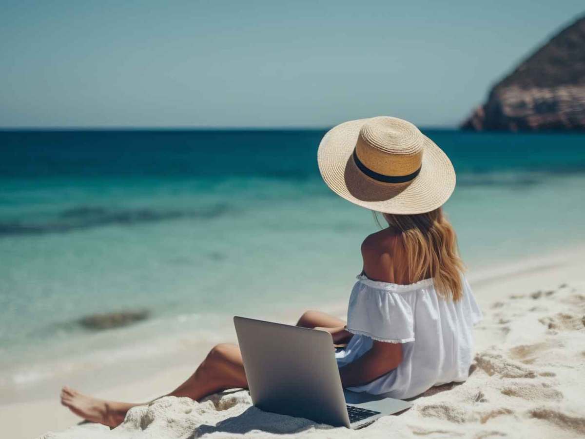 Junge Frau mit Sonnenhut sitzt am Strand mit ihrem Laptop und hat Urlaub.
