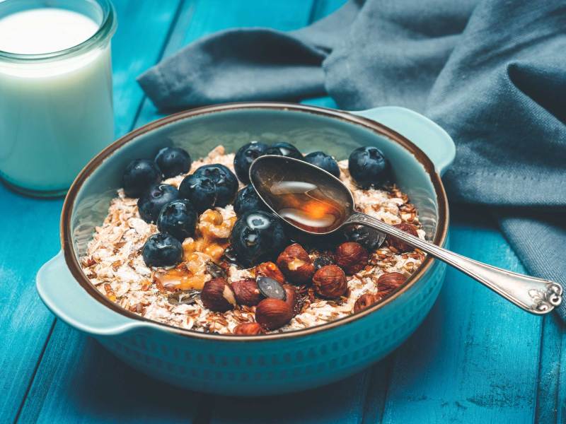 Blaubeeren und Nüsse auf Haferflocken in einer türkisfarbenen Schale, daneben ein Glas Milch.