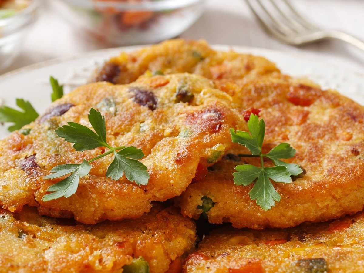 Mehrere der Tex-Mex-Kartoffelpuffer auf einem weißen Teller. Im Hintergrund unscharf geschnittenes Gemüse zu erkennen.