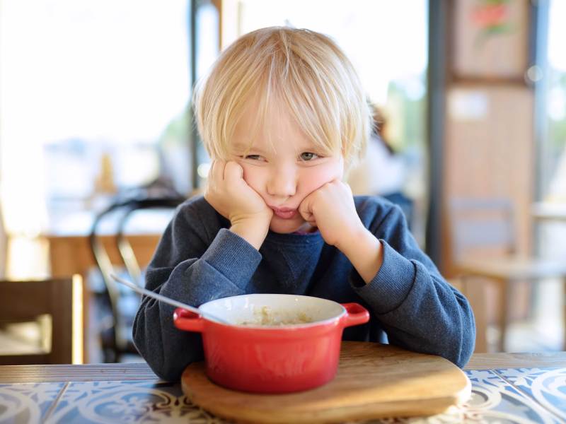 Kind sitzt bockig am Esstisch vor einer Schüssel und weigert sich zu essen.