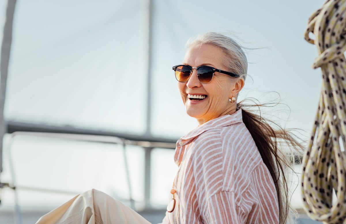 Frau über 50 sitzt draußen und trägt einen Pferdeschwanz und eine Sonnenbrille.