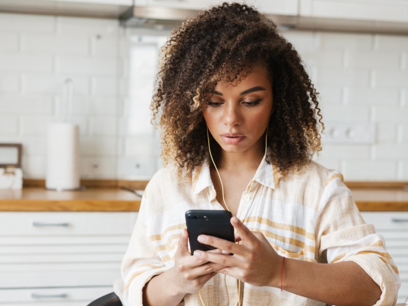 Frau mit ernstem Blick, die Kopfhörer in den Ohren hat und in der Küche sitzt, während sie auf skeptisch auf ihr Handy schaut