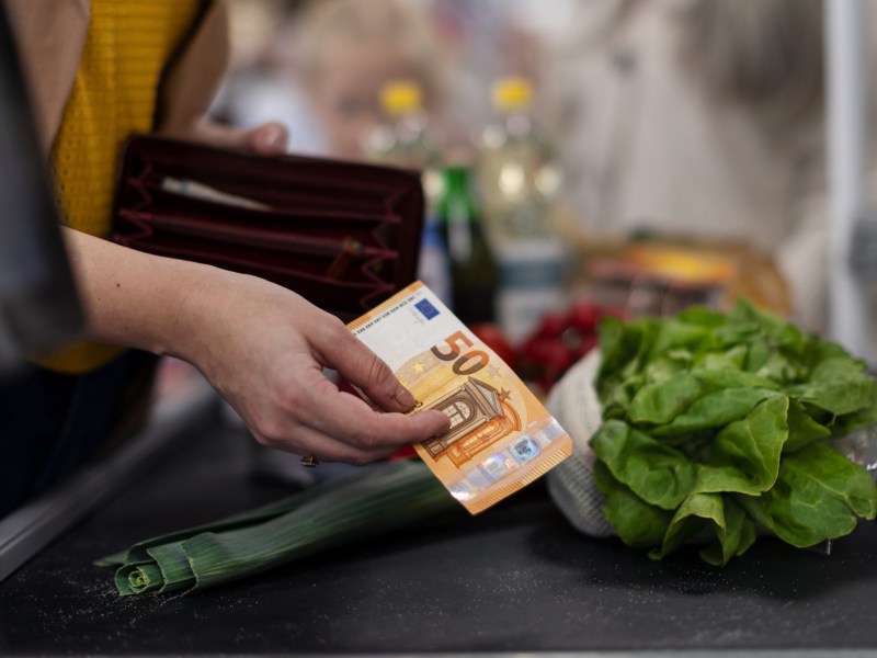 Frau, die einen 50-Euro-Schein in der Hand hält und auf dem Kassenband eine Gurke und einen Salatkopf gelegt hat