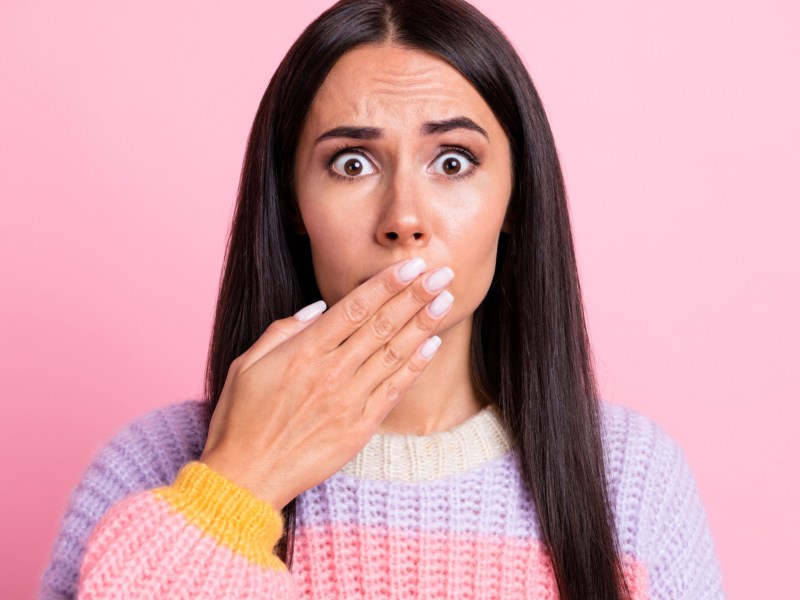 Frau in gestreiftem Pullover hält vor einem pinken Hintergrund ihre Hand vor den Mund, weil sie rülpsen muss.
