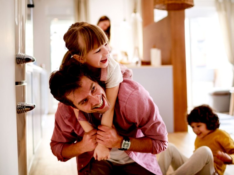 Vater tobt mit seinen beiden Kindern im Haus. Die Tochter trägt er gerade auf den Schultern, der Sohn sitzt im Hintergrund lachend auf dem Boden.