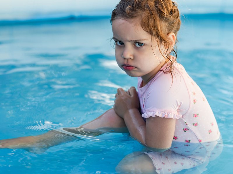 Mädchen, ca. drei Jahre, sitzt im Pool und schaut trotzig in die Kamera.