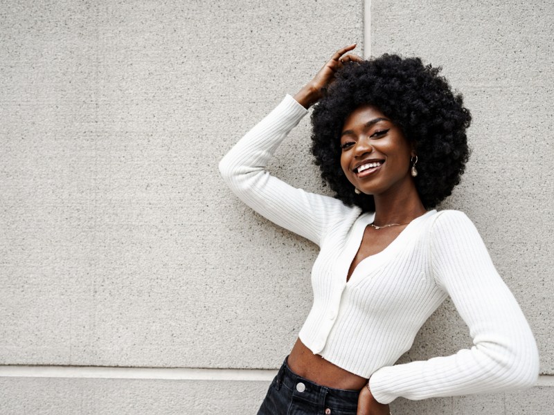 Frau mit stylishem Afro, die vor einer grauen Wand steht und in die Kamera lächelt