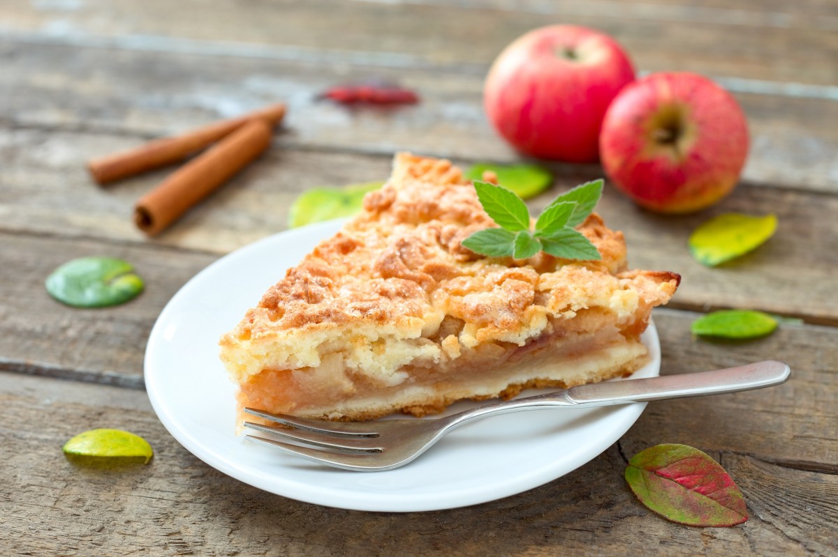 Ein Stück Apfelkuchen mit Zimtstreuseln mit einer Gabel auf einem Teller, garniert mit Minze, drumherum Äpfel und Zimtstangen.