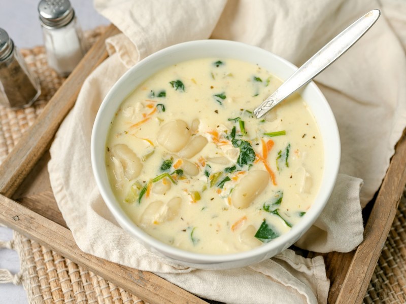 Eine weiße Suppenschale mit der Gnocchi-Suppe auf einem Holzbrett, welches wiederum auf einem weißen Tisch steht. Leicht schräg von oben fotografiert.