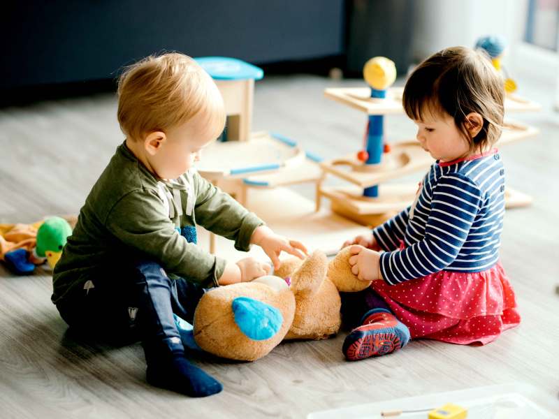 Zwei Kleinkinder sitzen auf dem Fußboden und spielen mit einem großen Plüschteddy.