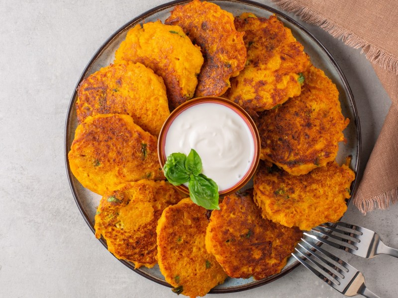 Ein dunkler Teller mit den Kartoffel-Kürbis-Puffern auf hell-grauem Untergrund direkt von oben fotografiert. Rechts unten im Bild ist noch ein braunes Tuch und zwei Gabeln sowie eine kleine Dip-Schale mit weißer Creme in der Mitte.