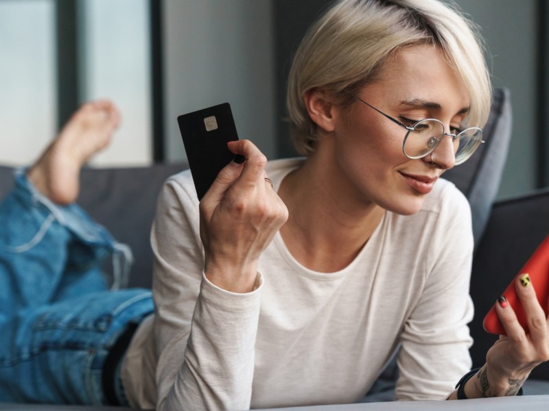 Frau mit kurzen Haaren liegt auf dem Sofa und hat Kreditkarte und Handy in der Hand.